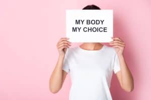 woman in white t-shirt holding placard with my body my choice lettering while covering face on pink