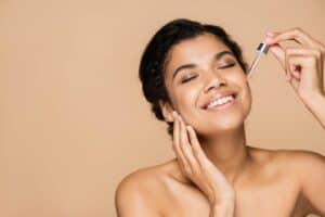 happy Blackwoman with eyes closed and bare shoulders applying serum with beige background